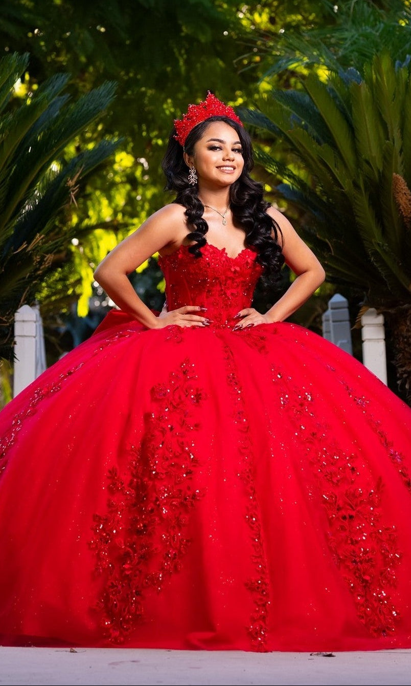 A young woman wearing the Quinceanera Dress CHF3129 By Chicas in vibrant red, featuring floral details and a matching crown, poses outdoors with a bright smile and her hands on her hips. The strapless ballgown is crafted from glitter tulle that shimmers in the light, set against a backdrop of lush green foliage.