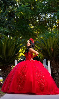 A young woman wearing the Quinceanera Dress CHF3129 By Chicas, a bright red embellished gown, and a tiara stands outdoors on a pathway surrounded by lush green plants and trees.