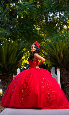 A young woman in the Quinceanera Dress CHF3129 By Chicas—a bright red, strapless ballgown adorned with a matching crown—stands outdoors, smiling amid lush greenery. The gown’s layers of glitter tulle shimmer beautifully in the natural light.