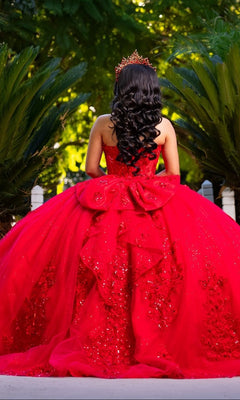 Wearing the Quinceanera Dress CHF3129 By Chicas, a person adorned with a crown and a large bow stands outdoors amidst green foliage, facing away.