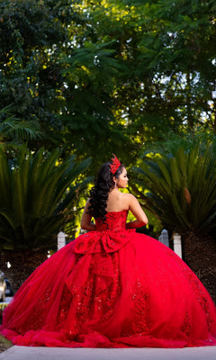 A young woman wearing the Quinceanera Dress CHF3129 By Chicas, featuring sparkling red glitter tulle and a matching crown, stands outdoors facing away, surrounded by green foliage.