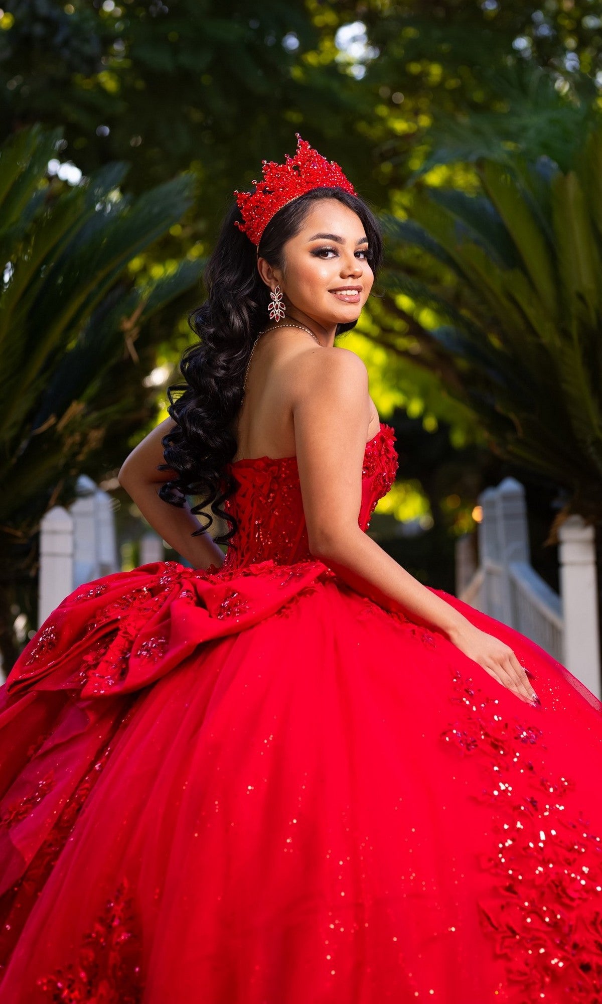 A young woman in the Quinceanera Dress CHF3129 By Chicas, a strapless ballgown in bright red with sparkling glitter tulle, poses outdoors on stone steps surrounded by greenery. Her elegant crown and the shimmering details of her gown add a magical touch to the scene.