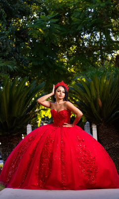A woman wearing the Quinceanera Dress CHF3129 by Chicas—a bright red, embellished gown with glitter tulle that catches the light—poses outdoors in front of lush green foliage, standing gracefully on a path with a crown atop her head, adding extra sparkle to her regal appearance.