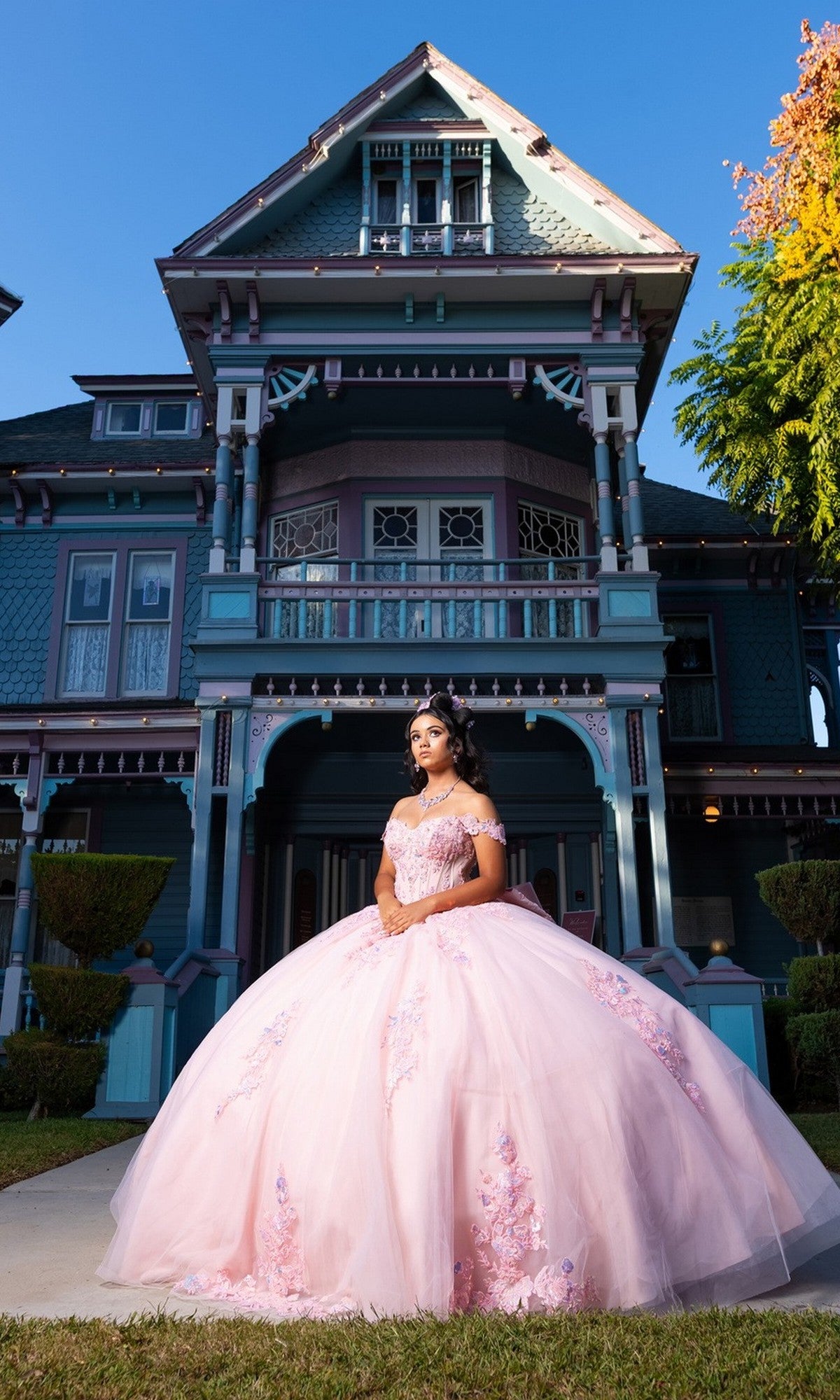 A young woman stands in front of a large, ornate Victorian-style house on a sunny day, wearing the Quinceanera Dress CHF3130 By Chicas—a pink off-the-shoulder gown featuring a delicate tulle ballgown skirt and corset-style bodice.