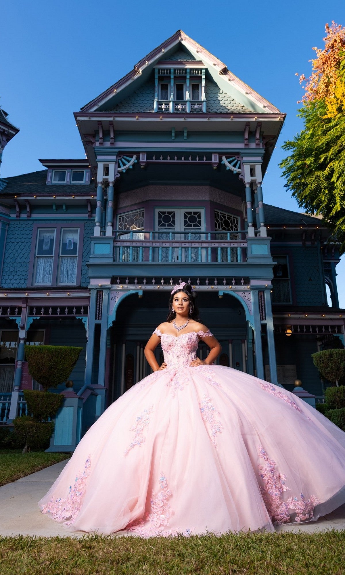 A young woman wearing the Quinceanera Dress CHF3130 By Chicas, featuring an off-the-shoulder design and a flowing tulle ballgown skirt, stands in front of a large, ornate Victorian-style house on a sunny day.