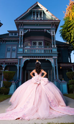Wearing the Quinceanera Dress CHF3130 by Chicas—an elegant, large pink ball gown with a corset-style bodice—a girl stands with her back to the camera, gazing at a grand Victorian house beneath a clear blue sky.