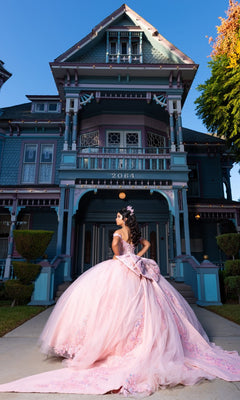 Wearing the Quinceanera Dress CHF3130 By Chicas—a pink, off-the-shoulder gown with a corset-style bodice—a woman stands in front of a grand Victorian-style house with blue and purple trim, glancing over her shoulder.