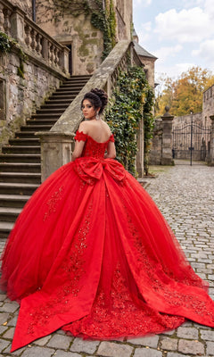 A woman stands on a cobblestone path near stone stairs and an ivy-covered building, wearing the Quinceanera Dress CHF3130 By Chicas—an ornate red ball gown with a tulle skirt, corset-style bodice, and a large bow.