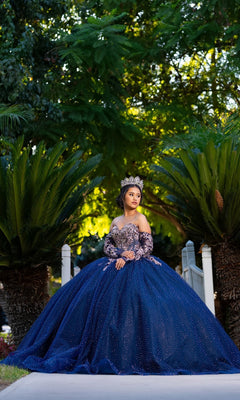 A young woman wearing the Quinceanera Dress CHF3131 By Chicas and a sparkling tiara sits on a bridge amidst lush green plants and trees, gazing off to the side.