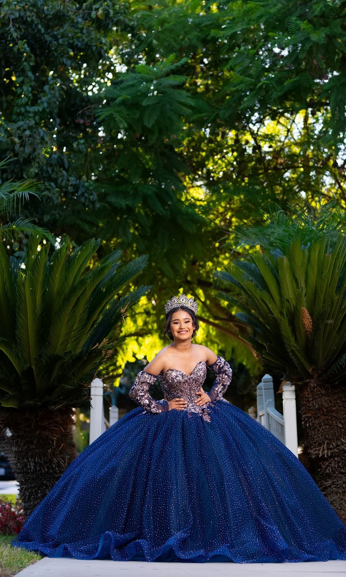 A young woman wearing the Quinceanera Dress CHF3131 By Chicas—a sparkly blue tulle ballgown paired with a crown—stands on a path between green palm trees, smiling at the camera.