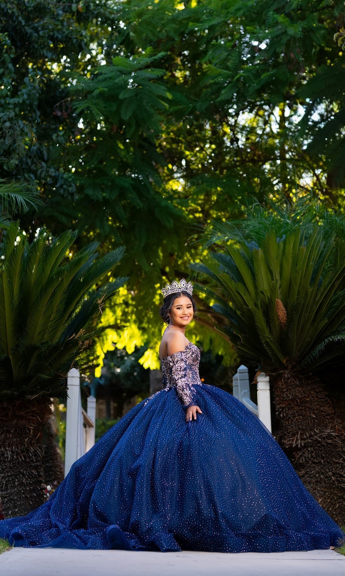 A young woman stands on a path between green bushes, looking back over her shoulder while wearing the sparkling blue Quinceanera Dress CHF3131 by Chicas and a crown.