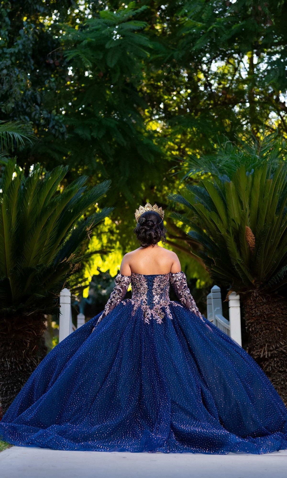A person in the Quinceanera Dress CHF3131 By Chicas—a dazzling navy, off-the-shoulder gown—stands outdoors between two large green plants, facing away from the camera. The shimmering fabric of the dress catches the light, adding an air of elegance and celebration to the scene.