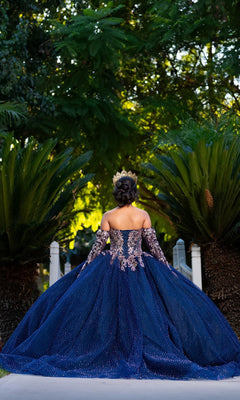 A person in the Quinceanera Dress CHF3131 By Chicas—a dazzling navy, off-the-shoulder gown—stands outdoors between two large green plants, facing away from the camera. The shimmering fabric of the dress catches the light, adding an air of elegance and celebration to the scene.