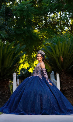 A young woman in the Quinceanera Dress CHF3131 By Chicas and a crown stands outdoors, smiling, with greenery and trees in the background.
