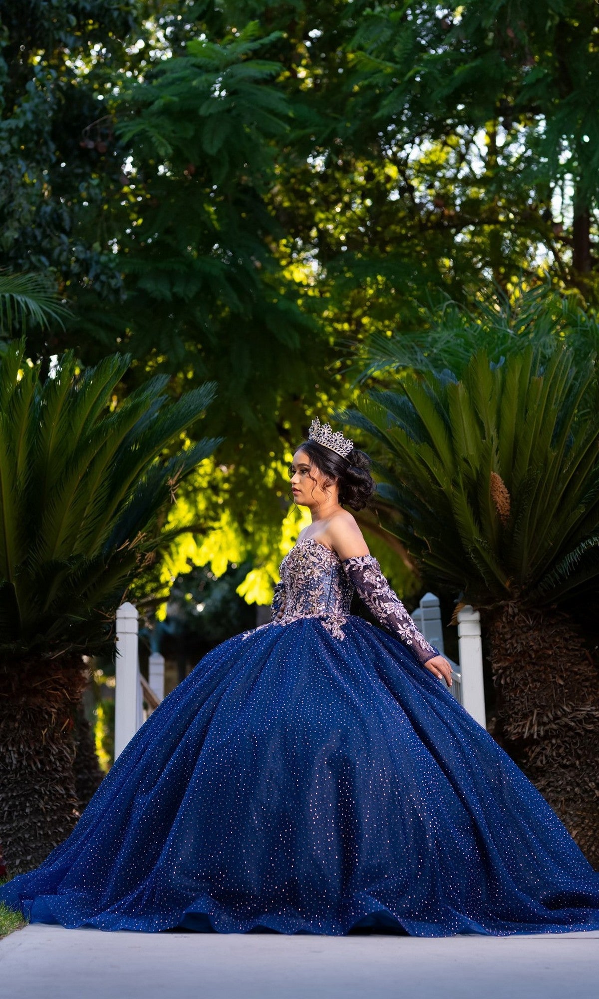 A young woman in the Quinceanera Dress CHF3131 By Chicas, a sparkling blue tulle ballgown paired with a tiara, stands outdoors on a path surrounded by lush green plants and trees.