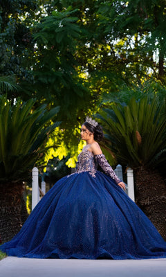 A young woman in the Quinceanera Dress CHF3131 By Chicas, a sparkling blue tulle ballgown paired with a tiara, stands outdoors on a path surrounded by lush green plants and trees.