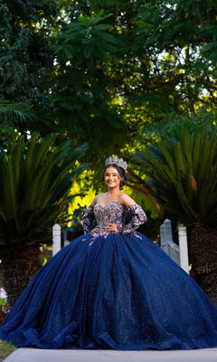 A young woman wearing the Quinceanera Dress CHF3131 By Chicas and a crown stands outdoors, smiling, with green foliage and trees in the background.