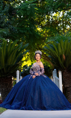 A young woman stands smiling on a path framed by palm trees and lush greenery, wearing the Quinceanera Dress CHF3131 By Chicas—a sparkling navy gown featuring a delicate tulle ballgown skirt—and a crown.