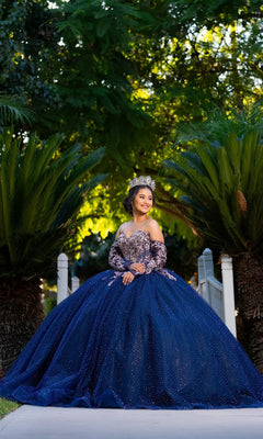 A young woman in the Quinceanera Dress CHF3131 By Chicas, a sparkling blue tulle ballgown, and crown stands on a path outdoors, surrounded by lush green trees and plants.