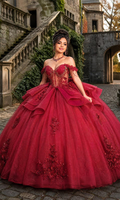 A woman in the Quinceanera Dress CHF3132 By Chicas, a stunning burgundy ballgown, stands on a cobblestone path near a stone staircase and an ivy-covered building. Her dress, adorned with delicate floral lace appliques, adds elegance and enchantment to the romantic setting.