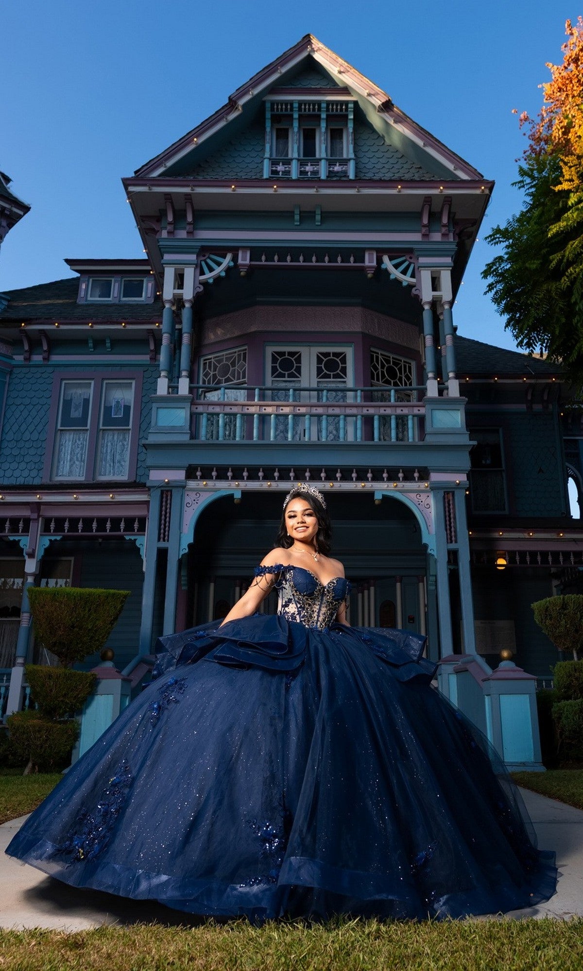 A young woman in the Quinceanera Dress CHF3132 by Chicas, a dark blue off-the-shoulder gown, stands smiling in front of a large, ornate Victorian-style house at dusk.