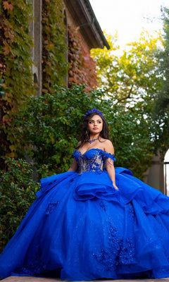 A young woman stands outdoors near a building draped in green vines, thoughtfully gazing to the side as her tiara glints in the sunlight. She wears the vibrant blue Quinceanera Dress CHF3132 By Chicas, featuring sparkling glitter-tulle and an elegant off-the-shoulder design.