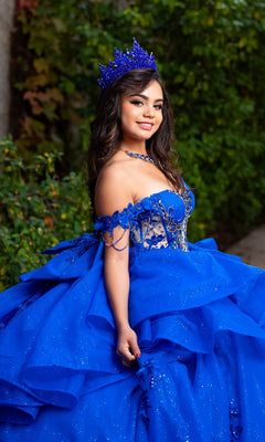 A young woman poses outdoors, smiling with greenery in the background, wearing the Quinceanera Dress CHF3132 by Chicas—a sparkling blue glitter-tulle ballgown with floral lace appliques and a matching crown.