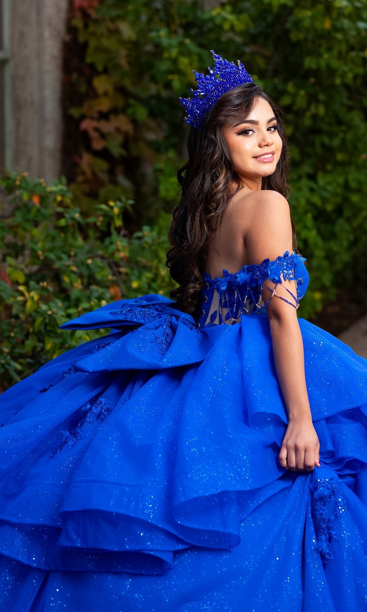 A woman stands outdoors among greenery, looking over her shoulder, wearing the Quinceanera Dress CHF3132 By Chicas—a bright blue glitter-tulle ballgown adorned with floral lace appliques and paired with a matching crown.