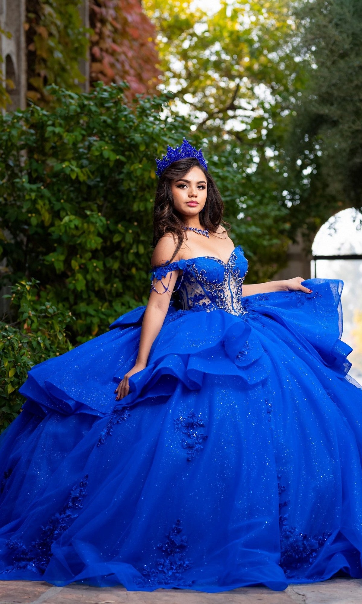A young woman poses outdoors against a lush, leafy backdrop wearing the Quinceanera Dress CHF3132 By Chicas—a vibrant blue off-the-shoulder gown featuring floral lace appliques and a matching crown.