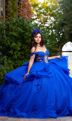 A young woman poses outdoors against a lush, leafy backdrop wearing the Quinceanera Dress CHF3132 By Chicas—a vibrant blue off-the-shoulder gown featuring floral lace appliques and a matching crown.