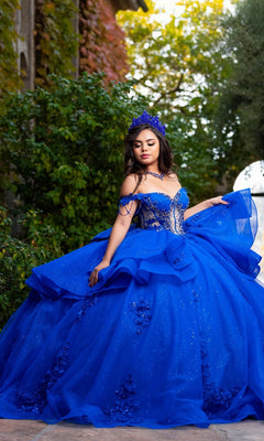 A young woman poses outdoors surrounded by greenery and sunlight, wearing the Quinceanera Dress CHF3132 By Chicas—a sparkly, bright blue off-the-shoulder gown adorned with delicate floral lace appliques and paired with a matching crown.