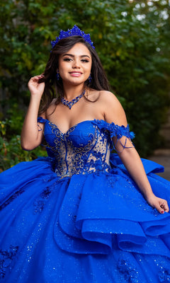 A young woman wearing the Quinceanera Dress CHF3132 By Chicas, a vibrant blue off-the-shoulder gown adorned with delicate floral lace appliques, stands outdoors in front of lush green foliage, smiling softly at the camera.