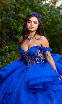 A young woman stands outdoors, surrounded by greenery, wearing the Quinceanera Dress CHF3132 By Chicas—a bright blue, ornate off-the-shoulder gown adorned with delicate floral lace appliques and matching accessories.