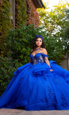 A young woman stands outdoors before lush green foliage and a building, wearing the Quinceanera Dress CHF3132 By Chicas—an elaborate, bright blue off-the-shoulder gown featuring floral lace appliques and delicate glitter tulle—while her crown catches the light.