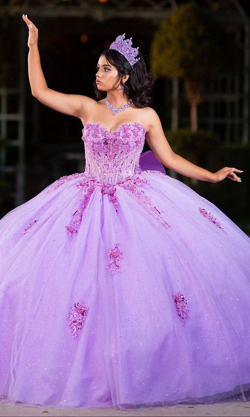 A young woman wearing the Quinceanera Dress CHF3133 By Chicas in lavender—a glitter tulle ballgown with a corset bodice—poses outdoors with one arm raised, accessorized with a tiara.