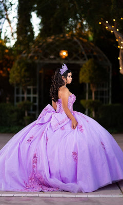 A young woman stands outdoors, facing sideways, wearing the Quinceanera Dress CHF3133 By Chicas—a lavender glitter tulle ballgown with a corset-style bodice and tiara—framed by trees and a gazebo in the background.