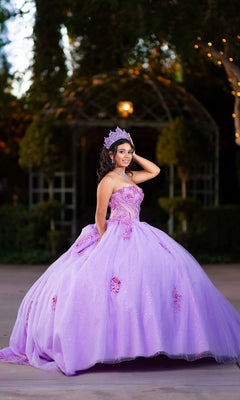 Wearing the Quinceanera Dress CHF3133 By Chicas, a young woman poses outdoors in a sparkling lavender glitter tulle ballgown and tiara, holding the side of her dress with trees and a gazebo in the background.