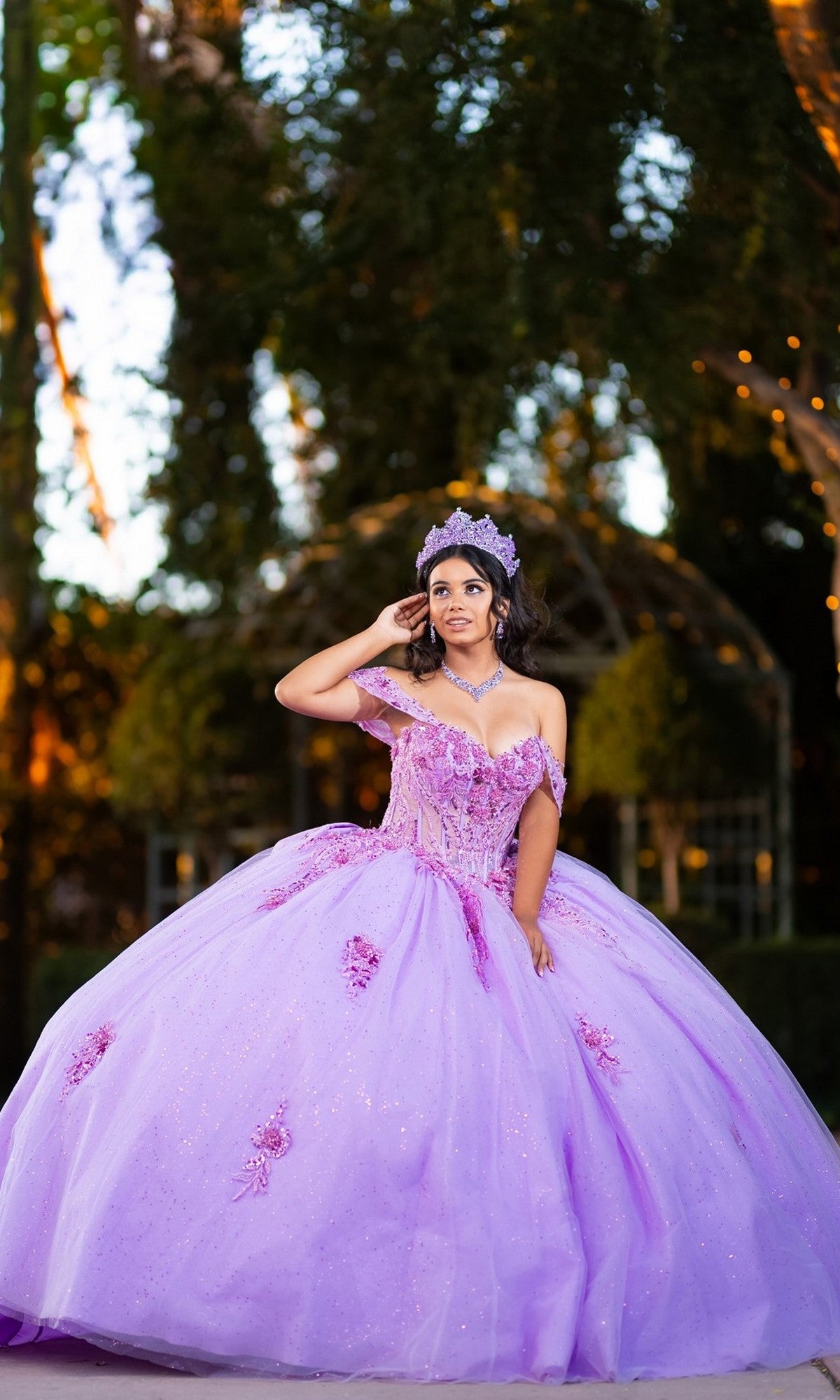 A young woman stands outdoors, posing for a formal photo with trees and lights in the background, wearing the Quinceanera Dress CHF3133 By Chicas—a large, ornate lavender strapless gown featuring a sparkling corset bodice and a glitter tulle ballgown skirt.