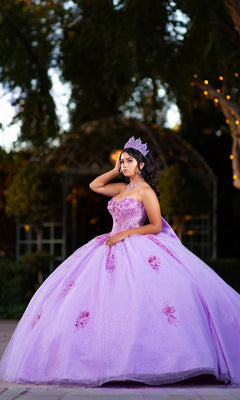 A young woman stands outdoors at dusk, posing with one hand to her hair while wearing the Quinceanera Dress CHF3133 By Chicas—a lavender glitter tulle ballgown with a corset-style bodice and crown—surrounded by trees and subtle string lights.