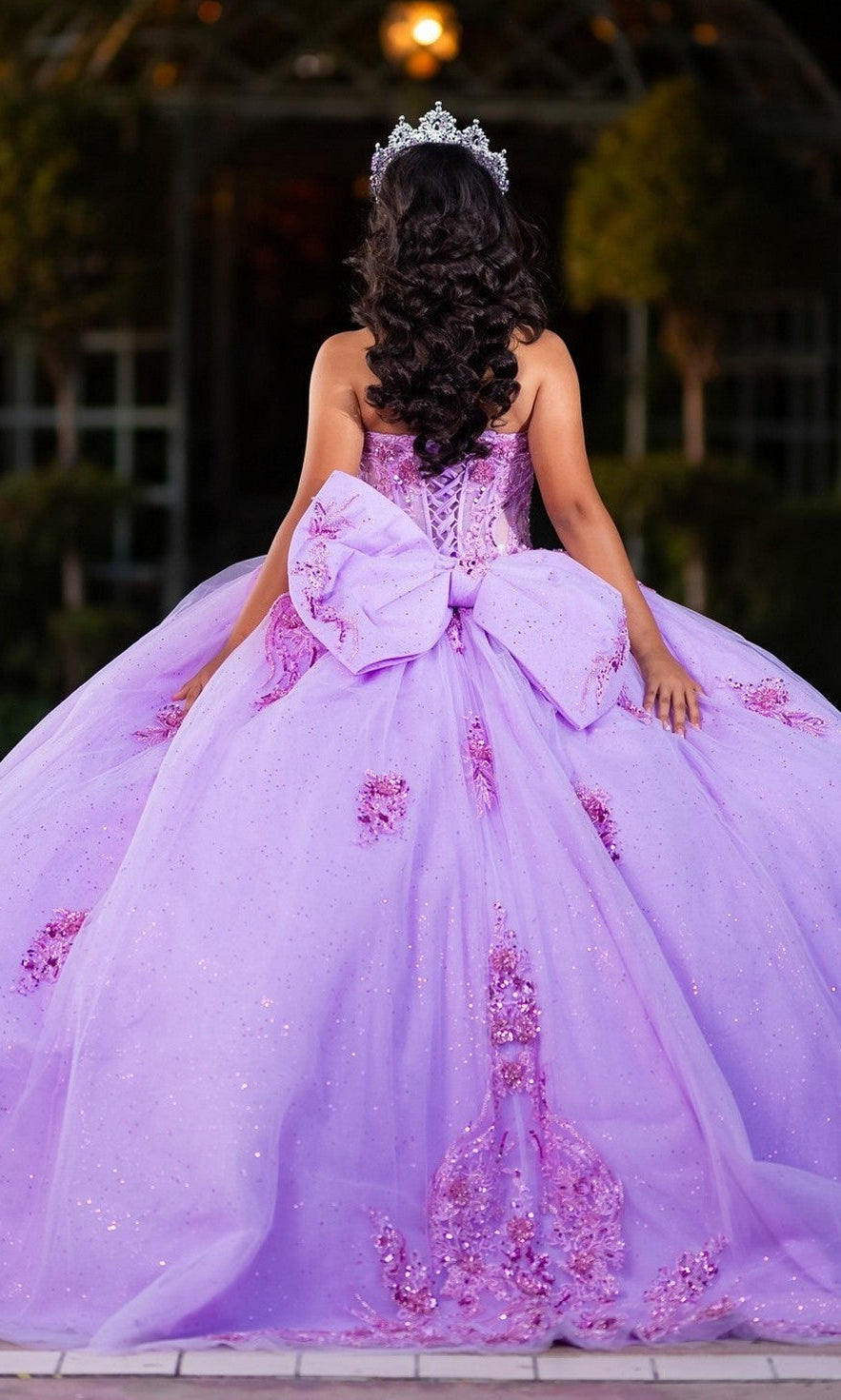 A girl stands outdoors, facing away from the camera, wearing the Quinceanera Dress CHF3133 By Chicas—a sparkling lavender glitter tulle ballgown adorned with a large bow and crowned with a tiara.