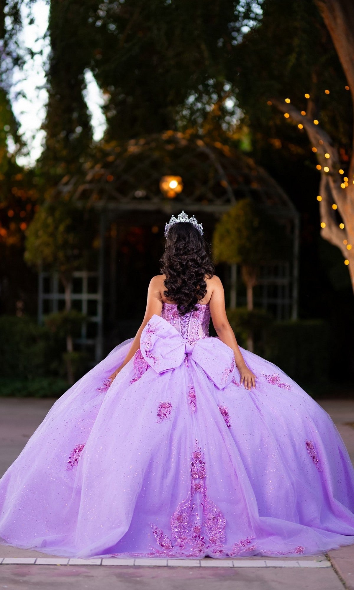 A young woman stands outdoors with her back to the camera, surrounded by trees and twinkling lights, wearing the Quinceanera Dress CHF3133 By Chicas—a lavender glitter tulle ballgown featuring a corset-style bodice and crowned with a tiara.