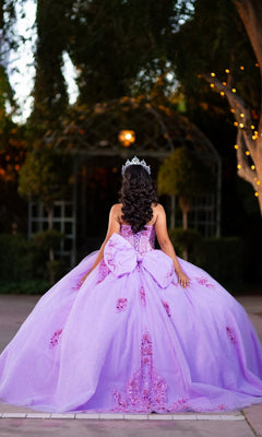 A young woman stands outdoors with her back to the camera, surrounded by trees and twinkling lights, wearing the Quinceanera Dress CHF3133 By Chicas—a lavender glitter tulle ballgown featuring a corset-style bodice and crowned with a tiara.