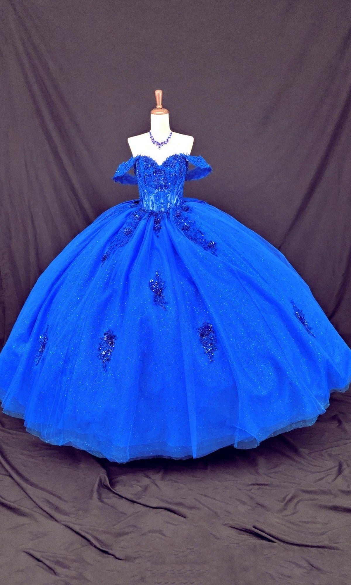 A mannequin displays the Quinceanera Dress CHF3133 By Chicas in royal blue, featuring off-the-shoulder sleeves, delicate floral appliqués, and a full tulle ballgown skirt—all set against a dark draped background.