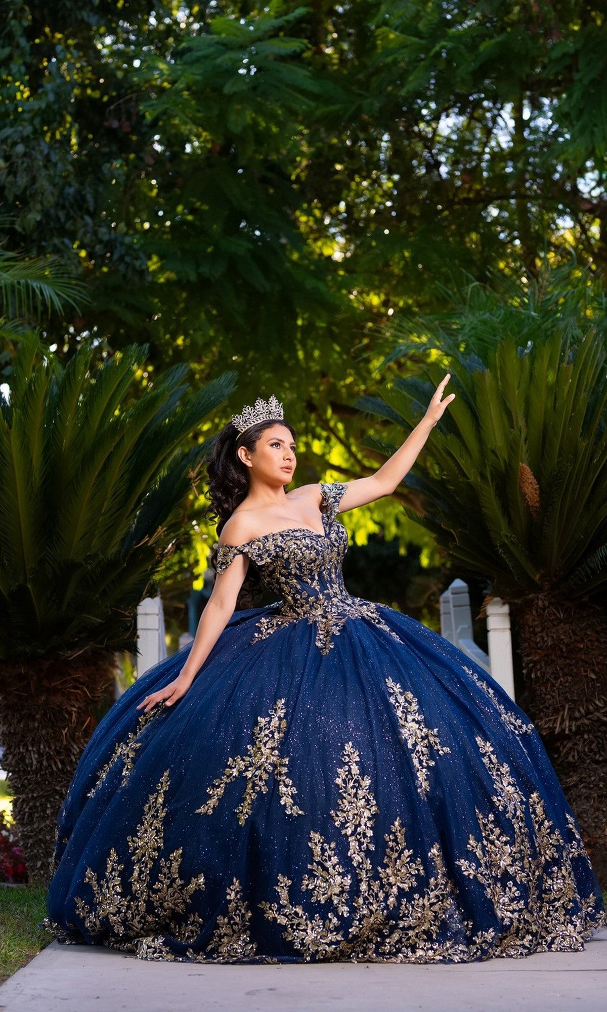 A woman in the Quinceanera Dress CHF3134 by Chicas, featuring an off-the-shoulder blue design with intricate gold embroidery and a crown, poses outdoors with one arm extended upward, surrounded by greenery.