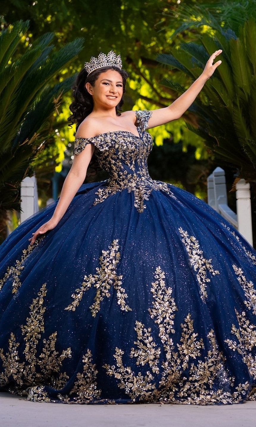 A young woman stands outdoors amid lush greenery, smiling and raising one arm while wearing the Quinceanera Dress CHF3134 by Chicas in Navy/Gold, adorned with gold embroidery and paired with a crown.