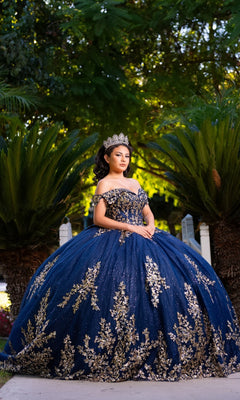 A young woman stands outdoors in front of lush green plants, wearing the Quinceanera Dress CHF3134 By Chicas—an off-the-shoulder blue and gold gown adorned with intricate gold embroidery—and accessorized with a tiara.