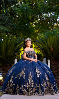 A young woman stands outdoors in front of green foliage, wearing the Quinceanera Dress CHF3134 By Chicas—an ornate off-the-shoulder blue and gold gown featuring intricate gold embroidery and a crown.