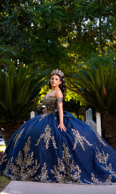 A young woman stands outdoors on a path, surrounded by lush green palm plants and trees, wearing the Quinceanera Dress CHF3134 By Chicas—a dark blue and gold gown featuring off-the-shoulder sleeves and delicate gold embroidery—looking at the camera.