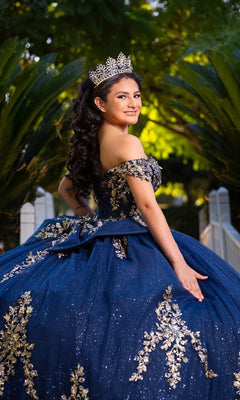 A young woman stands outdoors amid lush greenery, smiling and posing in the Quinceanera Dress CHF3134 By Chicas—a stunning blue and gold off-the-shoulder gown adorned with gold embroidery—while wearing a tiara.