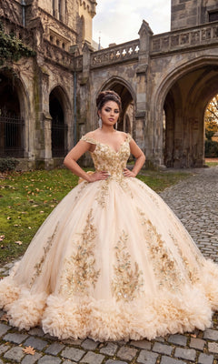 A woman wearing the Quinceanera Dress CHF3135 By Chicas in Champagne Gold, featuring ornate off-the-shoulder styling and elegant gold floral details, stands on a cobblestone path in front of a historic stone building adorned with arches and lush greenery. The lace-up back ballgown perfectly complements the timeless charm of her surroundings.